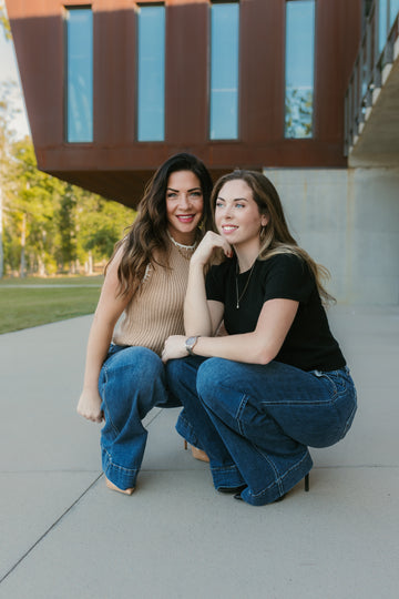 Two women sitting on a concrete surface with a modern building in the background. Stockbridge Styles clothing store