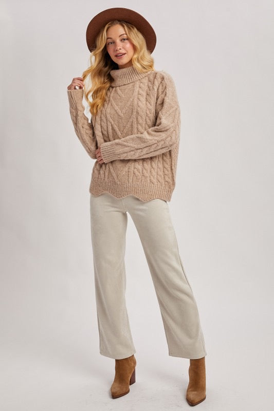 Woman wearing a beige cable knit sweater, white pants, and brown boots on a plain background
