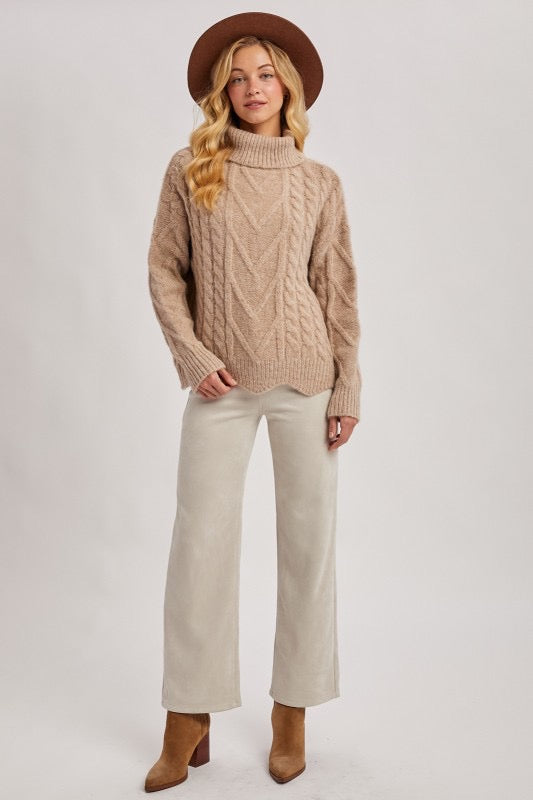 Woman wearing a beige cable knit sweater, white pants, and brown boots on a plain background