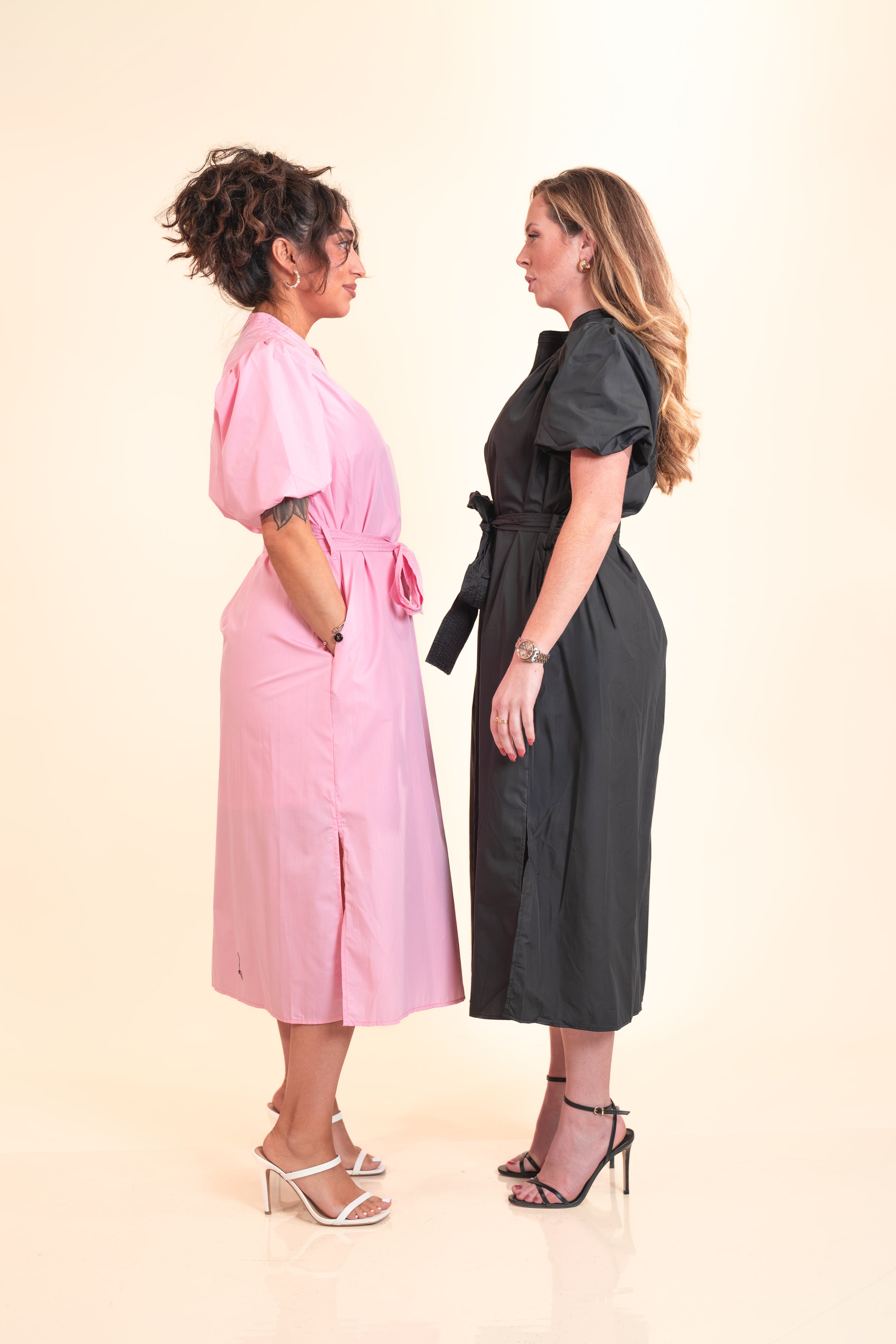 Two women wearing pink and black dresses standing back-to-back against a beige background