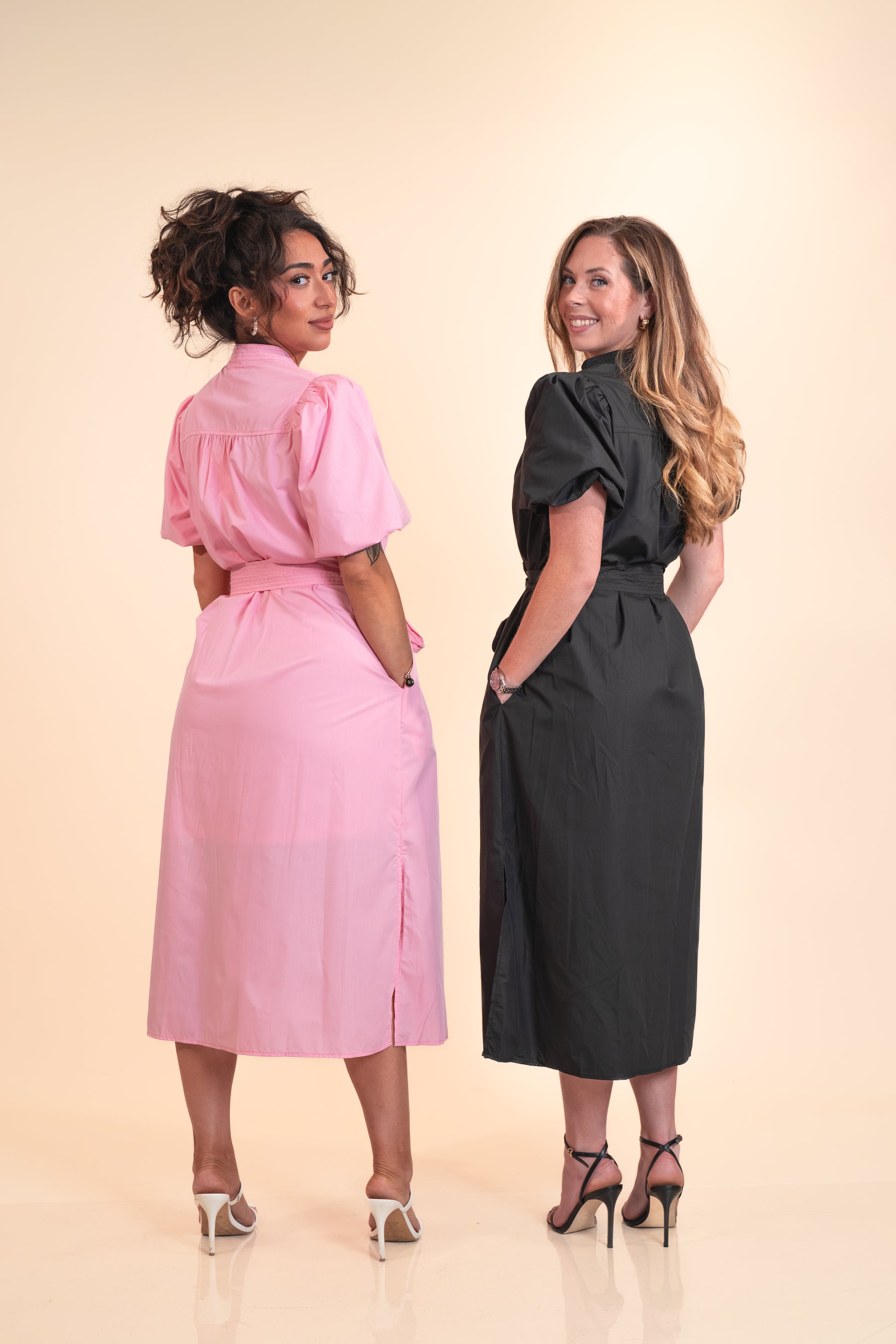 Two women wearing pink and black dresses standing back-to-back on a beige background