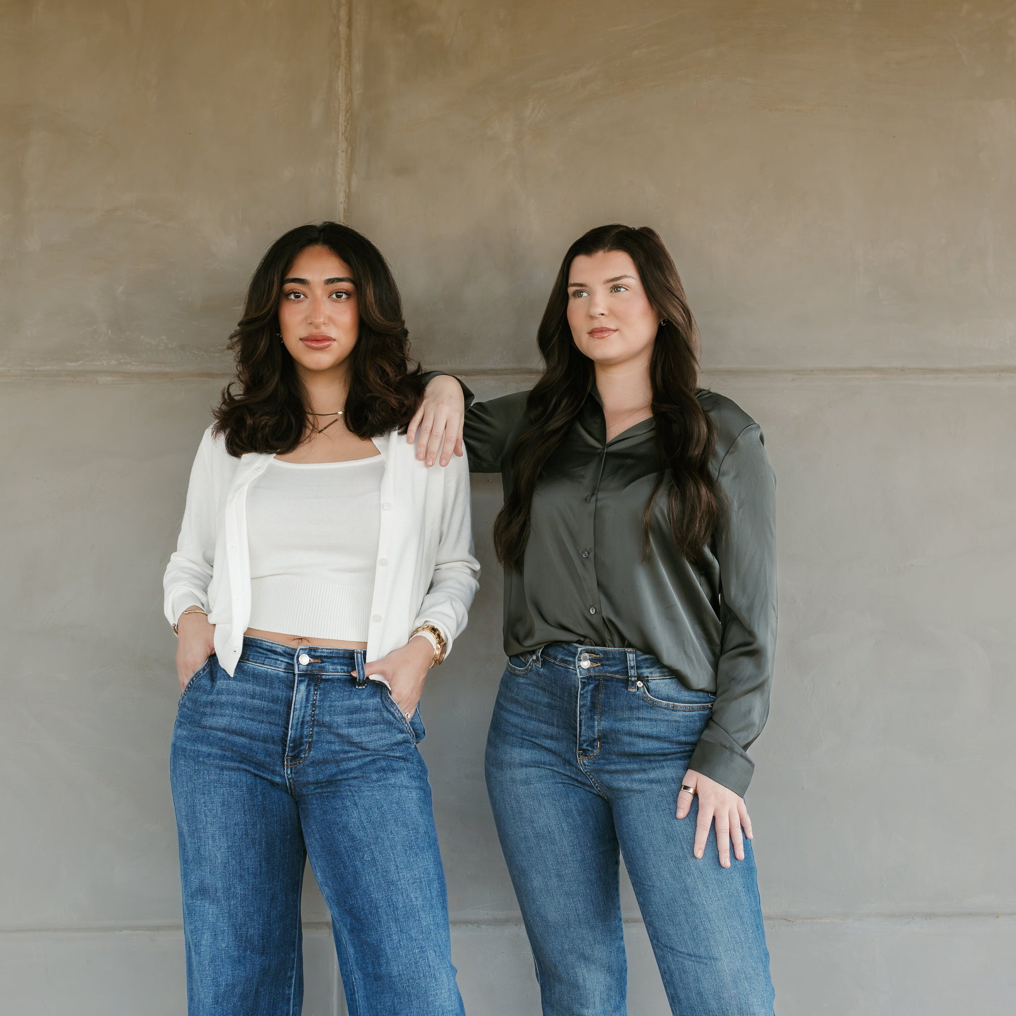 Two women wearing blue jeans standing against a plain wall. Stockbridge Styles just blue jeans. white sweater set with tank top. olive green satin top 