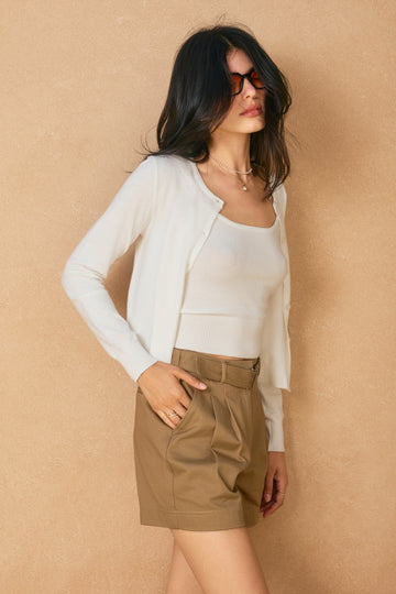 Woman wearing a white cardigan, white top, and brown shorts against a beige background